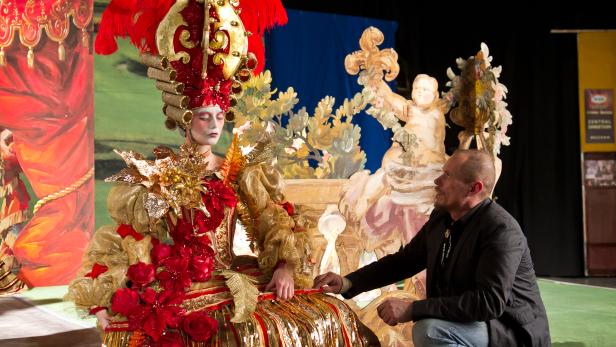 Eine Frau in einem aufwendigen, goldenen Kostüm mit roten Akzenten und Federn, neben einem Mann in Jeans.