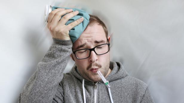 Ein Mann mit Brille und Fieberthermometer im Mund hält einen Eisbeutel an seinen Kopf.