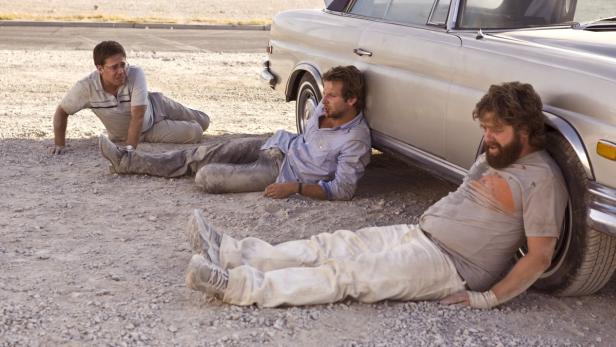 Bradley Cooper, Zach Galifianakis und Ed Helms liegen erschöpft in der Wüste neben einem Auto.