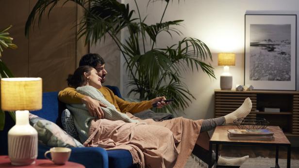 Ein Paar kuschelt sich auf einem blauen Sofa unter einer Decke und schaut fern.