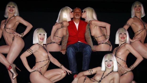 Ein Mann sitzt umgeben von mehreren leicht bekleideten Frauen mit blonden Perücken.