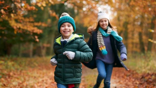 Zwei Kinder rennen lachend durch einen herbstlichen Wald.