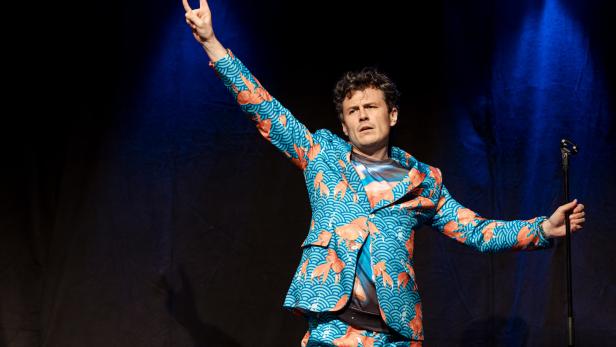 Ein Mann in einem Anzug mit Goldfischmuster steht auf einer Bühne und gestikuliert.