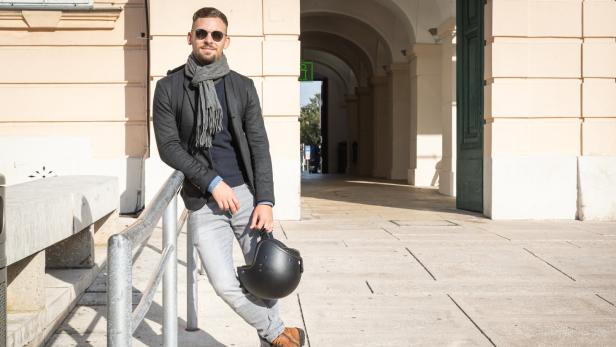 Ein Mann mit Sonnenbrille lehnt an einem Geländer und hält einen Motorradhelm.