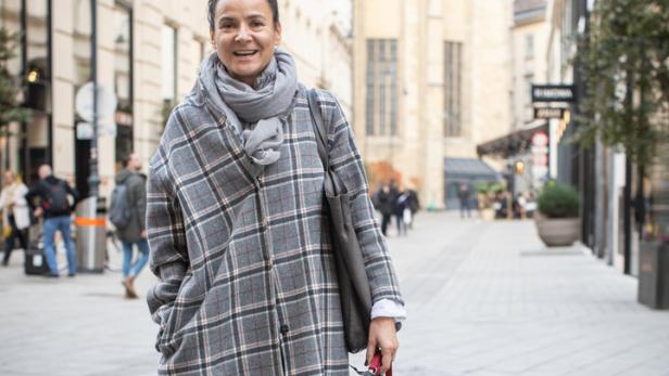 Eine Frau mit einem karierten Mantel und einem Schal geht mit ihrem Hund in einer Stadt spazieren.