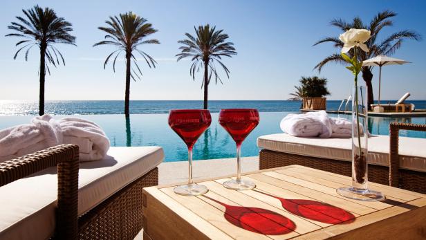 Zwei rote Weingläser stehen auf einem Tisch an einem Pool mit Blick auf das Meer und Palmen.