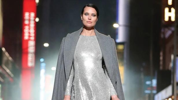 Eine Frau in einem silbernen Kleid und einem grauen Mantel geht barfuß auf einer Straße in New York.