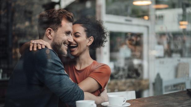Ein lachendes Paar umarmt sich in einem Café bei zwei Tassen Kaffee.