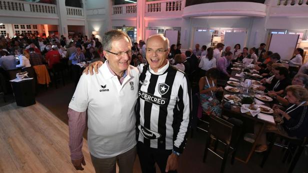 Zwei Männer posieren auf einer Veranstaltung mit vielen Gästen im Hintergrund.