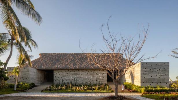 Ein Haus mit einem Strohdach und einer Fassade aus Beton befindet sich in einer tropischen Landschaft.