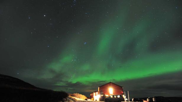 Grüne Nordlichter über einem roten Haus mit geparkten Autos.