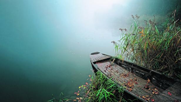 Ein verlassenes Holzboot liegt am Ufer eines nebligen Sees.