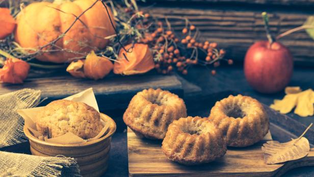 Ein Kürbis, Lampions, ein Apfel und kleine Gugelhupfe auf einem Holztisch.