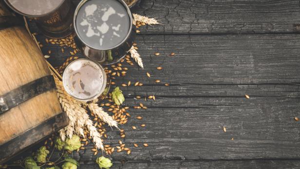 Drei Gläser Bier, Gerste, Hopfen und ein kleines Fass auf einem dunklen Holztisch.