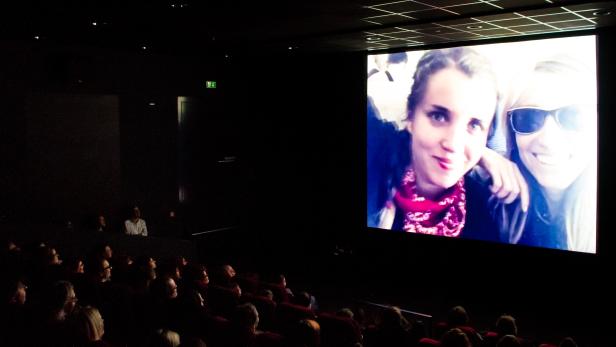 Zuschauer sitzen in einem dunklen Kinosaal vor einer großen Leinwand, auf der ein Selfie von zwei Frauen zu sehen ist.