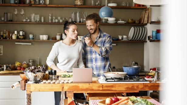 Ein Paar kocht gemeinsam in einer Küche, wobei der Mann die Frau mit Pasta füttert.