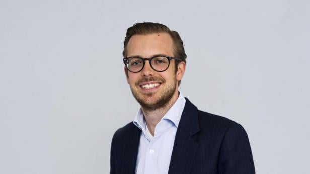 Ein lächelnder Mann mit Brille und Sakko vor einem hellgrauen Hintergrund.