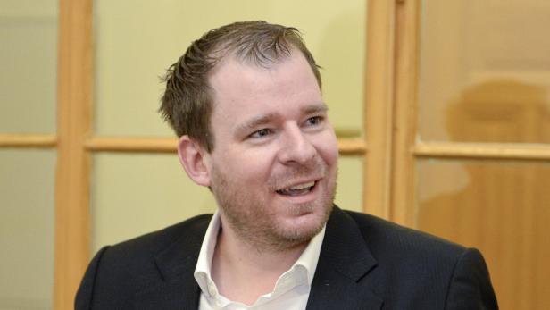 Ein Mann in einem dunklen Anzug und einem weißen Hemd lächelt.