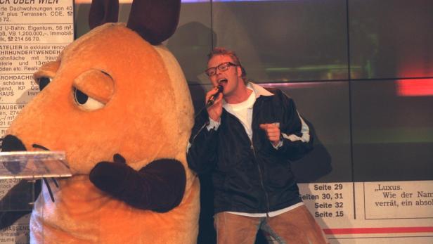 Ein Mann mit Brille singt neben einer überdimensionalen „Maus“-Figur in ein Mikrofon.