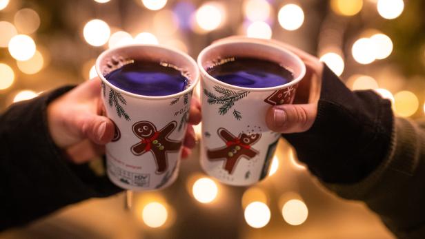 Zwei Hände halten Becher mit Glühwein vor einem Hintergrund mit Lichtern.