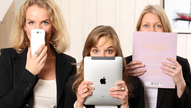 Drei Frauen halten ein Smartphone, ein Tablet und ein Buch vor ihre Gesichter.