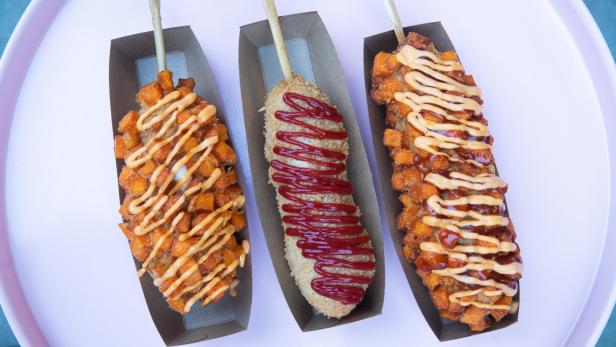 Drei koreanische Corn Dogs mit verschiedenen Toppings auf einem rosa Tablett.
