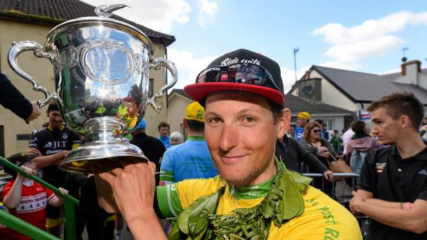 Ein Radrennfahrer hält einen silbernen Pokal und trägt einen Lorbeerkranz.