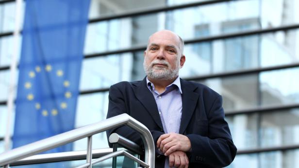 Ein Mann mit Bart lehnt vor einem Gebäude mit der Europaflagge im Hintergrund.