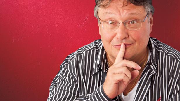 Ein Mann mit Brille hält den Finger vor den Mund, um Stille zu signalisieren.