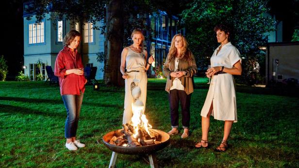 Vier Frauen stehen abends mit Getränken um eine Feuerschale im Garten.