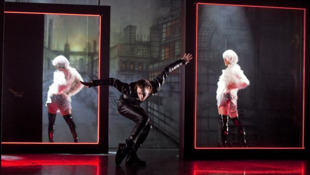 Ein Mann in schwarzer Lederkleidung tanzt zwischen zwei Frauen in weißen Kostümen auf einer Bühne.