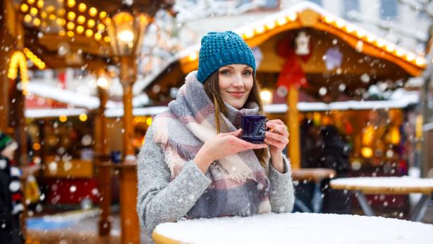 Eine Frau mit Mütze und Schal hält eine Tasse auf einem Weihnachtsmarkt im Schnee.