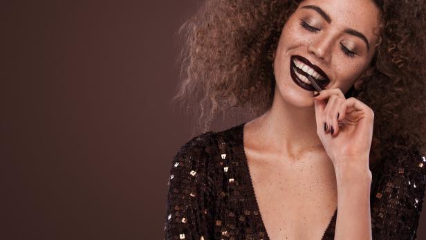 Eine Frau mit lockigem Haar und dunklem Lippenstift lächelt und hält ein Stück Schokolade zwischen den Zähnen.