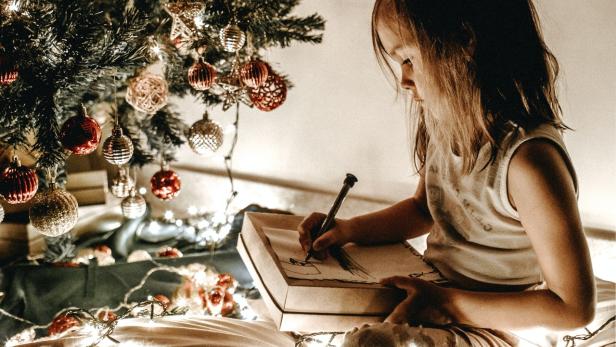 Ein Mädchen sitzt unter einem Weihnachtsbaum und malt in ein Buch.