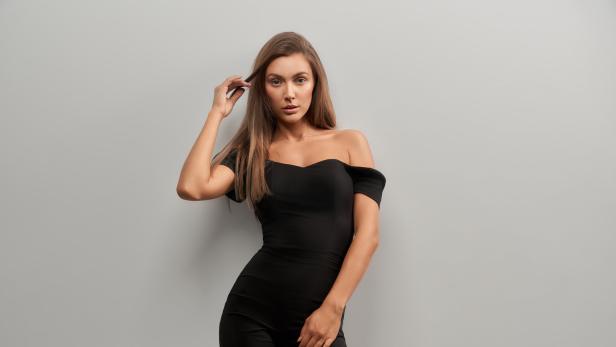 Eine Frau mit langen braunen Haaren posiert in einem schwarzen Overall vor einer grauen Wand.