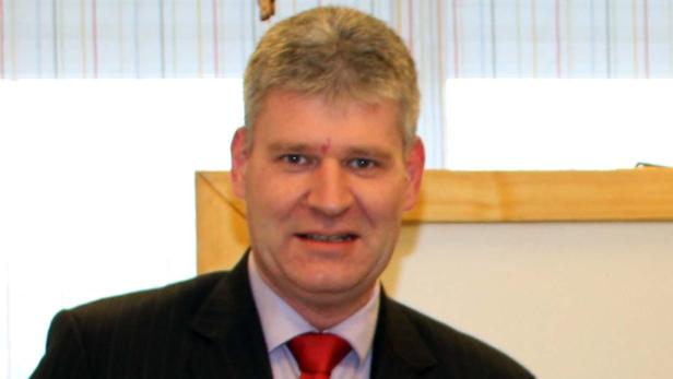 Ein Mann mit blonden Haaren, Anzug und roter Krawatte lächelt in die Kamera.