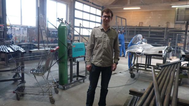 Ein Mann mit Brille steht in einer Werkstatt mit Schweißgeräten und Metallarbeiten.