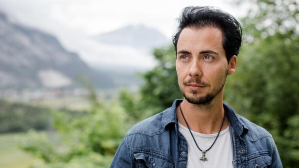 Ein Mann mit Bart und Jeansjacke blickt vor einer Bergkulisse in die Ferne.