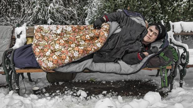 Ein Mann schläft im Winter auf einer Parkbank unter einer Decke.