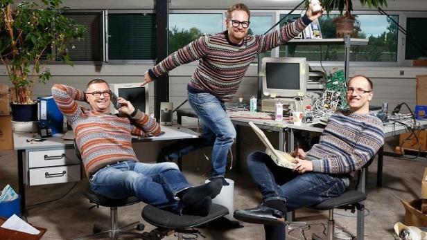Drei Männer sitzen und stehen an Schreibtischen voller Computer und Elektronik.