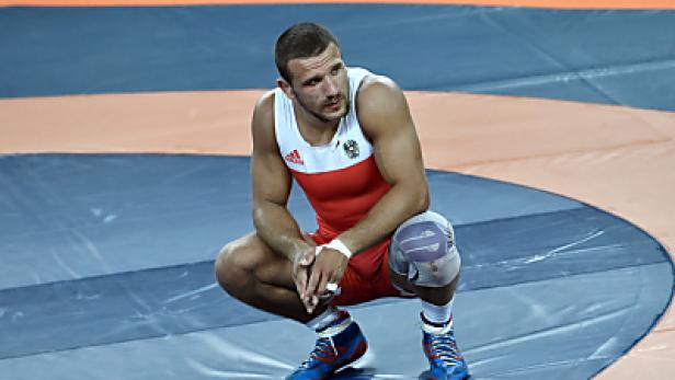 Ein Ringer in rot-weißer Kleidung hockt auf einer Ringermatte.