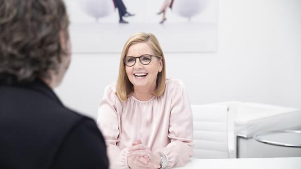 Eine lachende Frau mit Brille sitzt an einem Tisch und spricht mit einer Person.