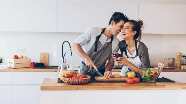 Ein lachendes Paar bereitet gemeinsam in einer hellen Küche eine Mahlzeit zu.