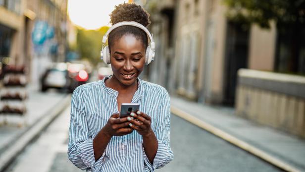 Eine junge Frau mit Kopfhörern schaut auf ihr Smartphone auf einer Straße.