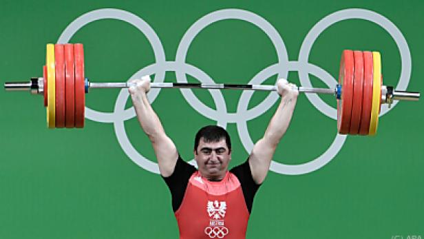 Ein Gewichtheber stemmt eine Langhantel vor dem Hintergrund der olympischen Ringe.