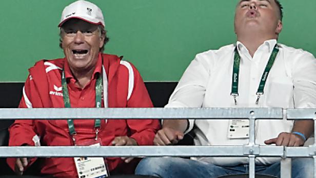 Zwei Männer sitzen auf einer Tribüne, der linke Mann jubelt.