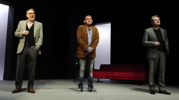 Drei Männer stehen auf einer Bühne vor einer roten Couch.