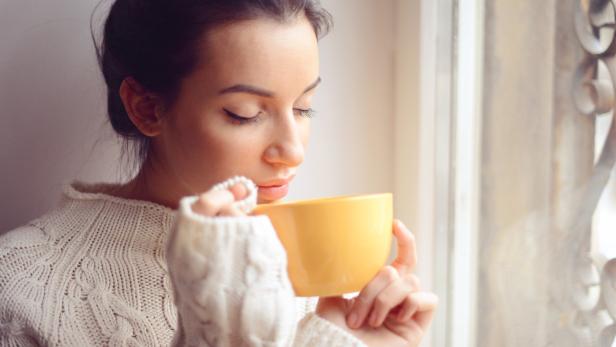 Eine Frau in einem weißen Pullover trinkt aus einer gelben Tasse am Fenster.