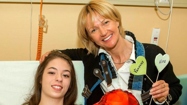 Eine Frau besucht ein junges Mädchen im Krankenhaus und hält Schilder mit den Namen „Vanessa“ und „Kira“.