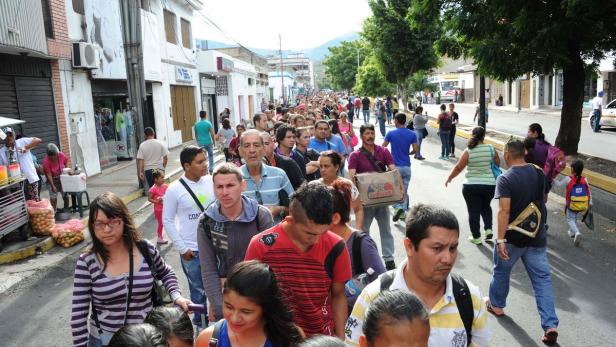 Eine lange Menschenschlange erstreckt sich eine Straße entlang, gesäumt von Gebäuden und Bäumen.